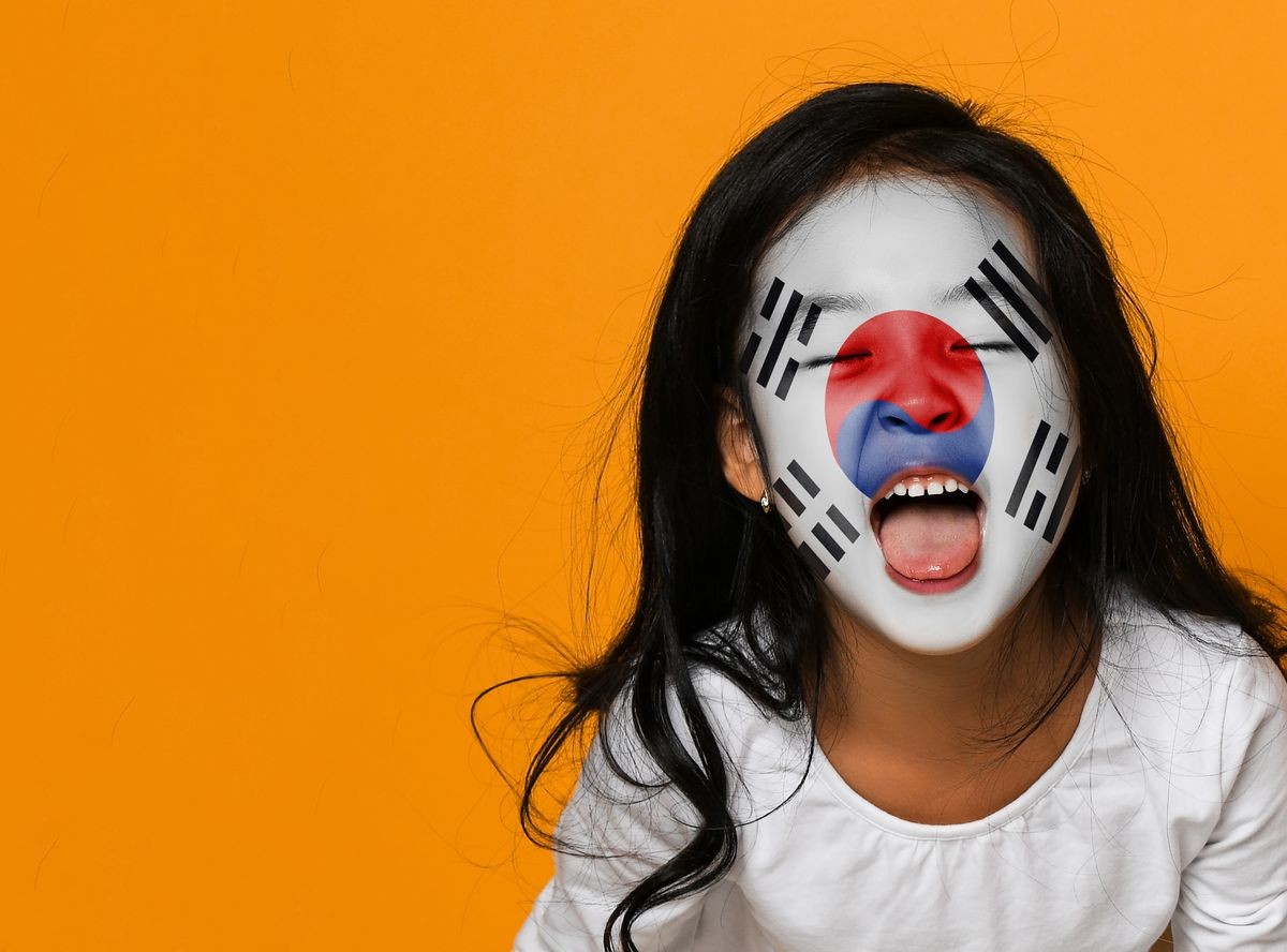 Portrait of cute little asian girl with a painted korean flag shows tongue over yellow wall background